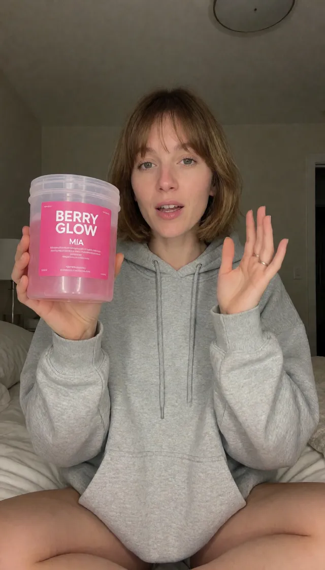 Mia, a fictional AI character, in a grey hoodie sitting on her bed holding a pink skincare tub